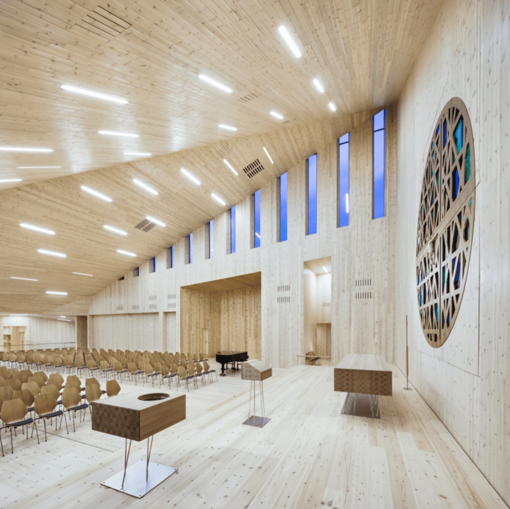 Modern light-wood church sanctuary with vaulted ceiling, stained glass window, and rows of seating.