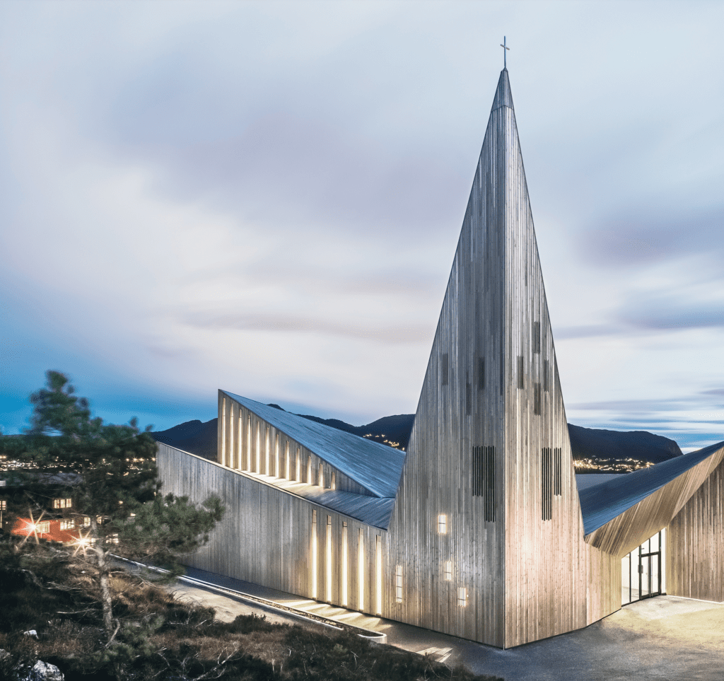 Contemporary church with dramatic angular architecture illuminated at dusk against a mountain backdrop.