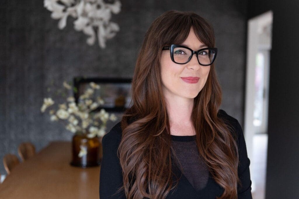 A professional waist-up portrait of a woman with long, wavy brown hair and bangs. She is wearing thick black-rimmed glasses, a subtle pink lipstick, and a black top, smiling warmly at the camera. The background is a softly blurred, moody dining space with a dark wall, a wooden table, and a vase with white flowers.
