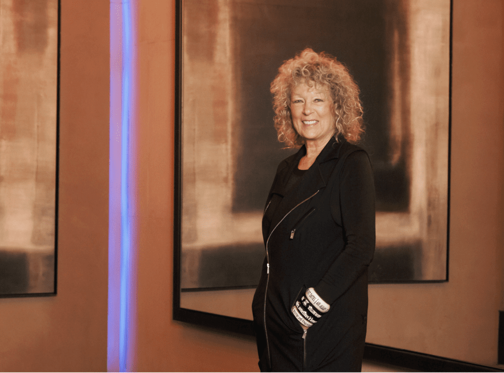 A portrait of a woman with short, curly blonde hair smiling, wearing a sleeveless black zip-up vest over a black top. She is standing with her hands in her pockets in front of large, framed abstract square artwork in neutral, earthy tones. A thin vertical blue neon light strips down a wall section to her left.