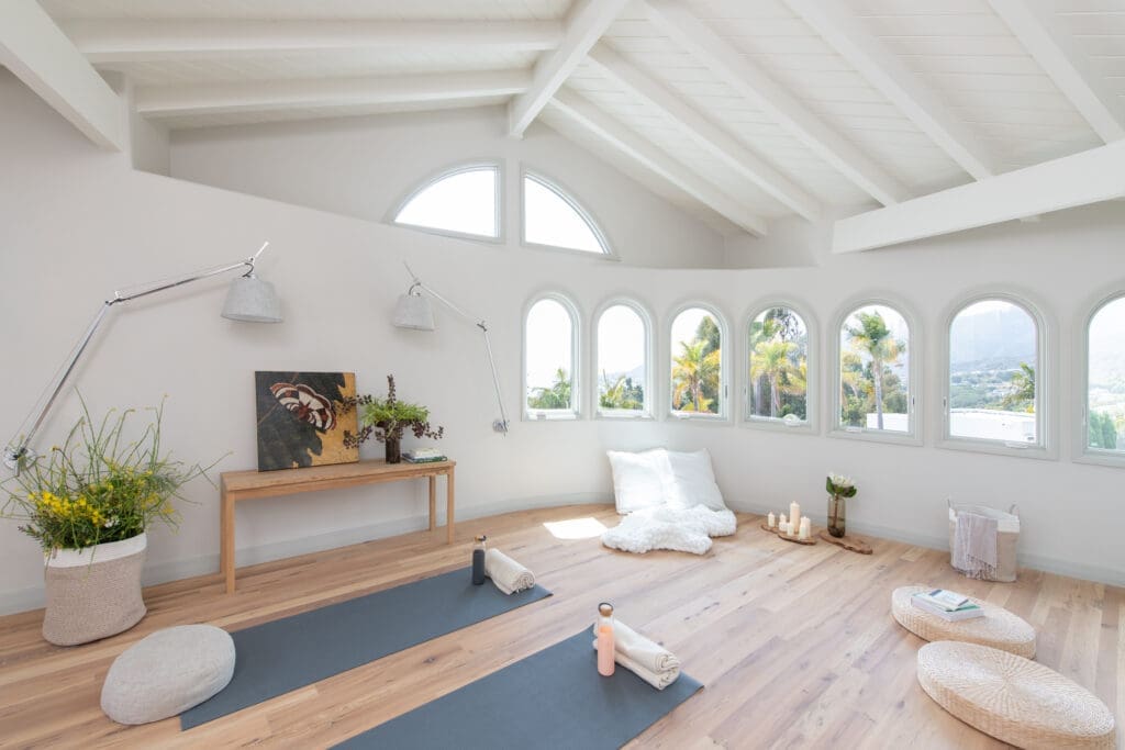 A bright, sunlit yoga and meditation studio with vaulted white ceilings, exposed beams, and light hardwood floors. Two dark gray yoga mats are laid out with rolled-up white towels and water bottles. A curved wall features a row of arched windows looking out at tropical palm trees. A simple wooden bench, a large basket filled with yellow wildflowers, and woven meditation cushions complete the minimalist, peaceful space.