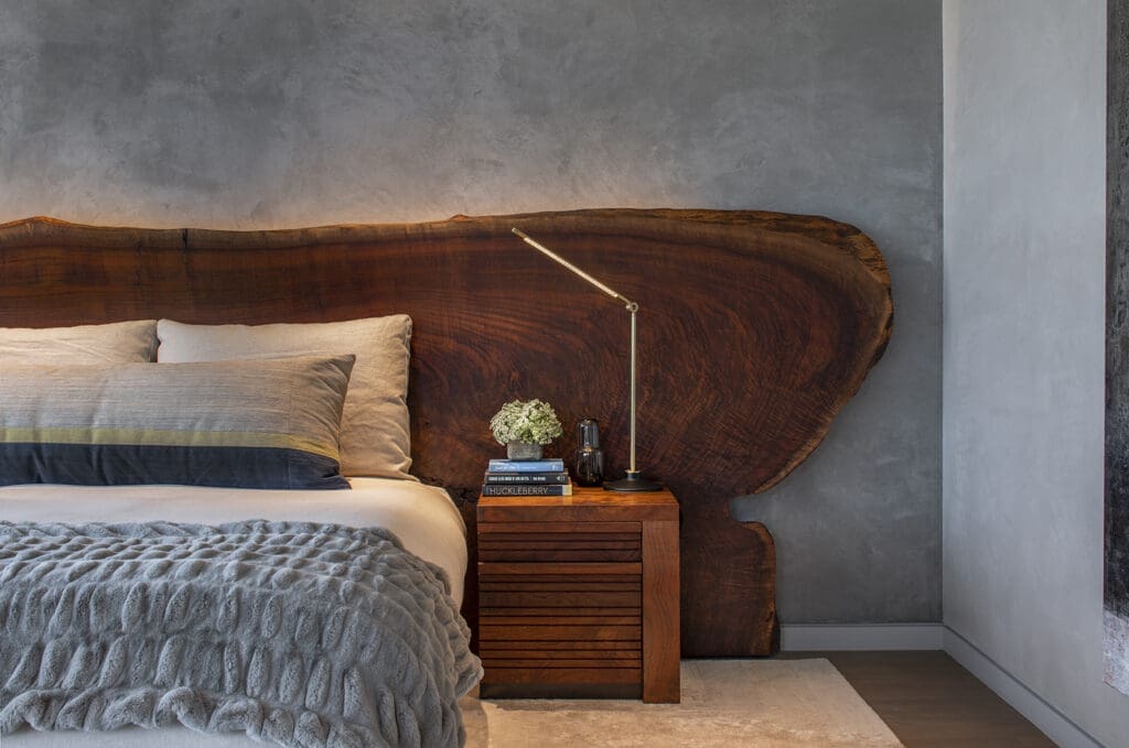 A close-up view of a modern platform bed featuring a massive, rustic live-edge dark wood slab headboard set against a textured gray concrete wall. The bed is dressed with a textured gray blanket and neutral-toned pillows. Next to the bed is a matching wood-slat nightstand holding a sleek, minimalist brass reading lamp and a small arrangement of white flowers.