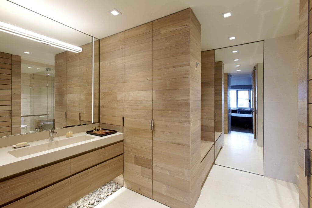 A contemporary, minimalist bathroom and dressing corridor clad entirely in light-toned natural wood paneling. A long white trough sink sits on a floating wooden vanity, highlighted by a bed of illuminated white river stones underneath. A large frameless mirror stretches across the wall, and a full-length mirrored panel at the end of the walkway reflects the clean, architectural lines of the space.