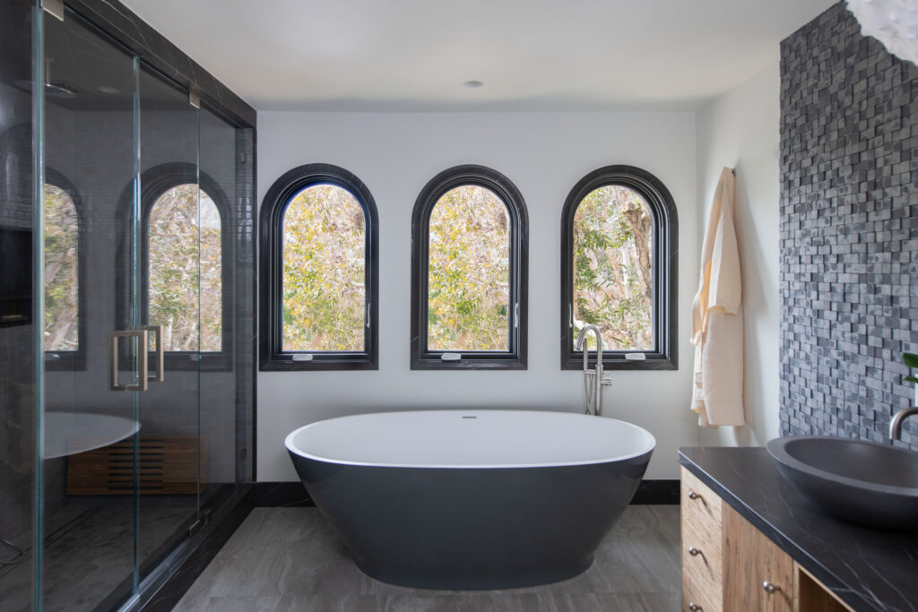 A modern primary bathroom featuring a dark charcoal-gray freestanding soaking tub centered below three matching arched windows with black frames. On the left, a spacious walk-in shower is enclosed in glass with dark gray tiling. On the right, a textured accent wall made of small, dark gray square tiles stands behind a floating wooden vanity with a dark vessel sink and a black marble countertop.