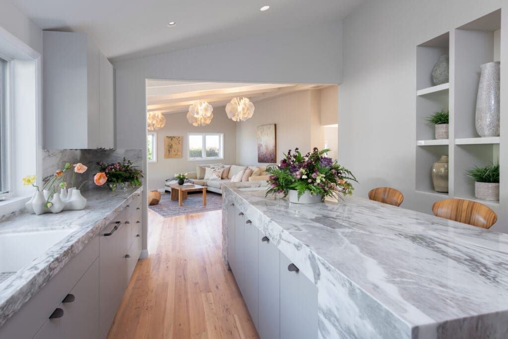 An open-concept kitchen looking into a bright living room. The kitchen features a long island with a striking white and gray veined marble waterfall countertop and light gray cabinetry. In the background, the living room is styled with a cream-colored sectional sofa, a wooden coffee table, and sculptural white floral pendant lights hanging from the ceiling.