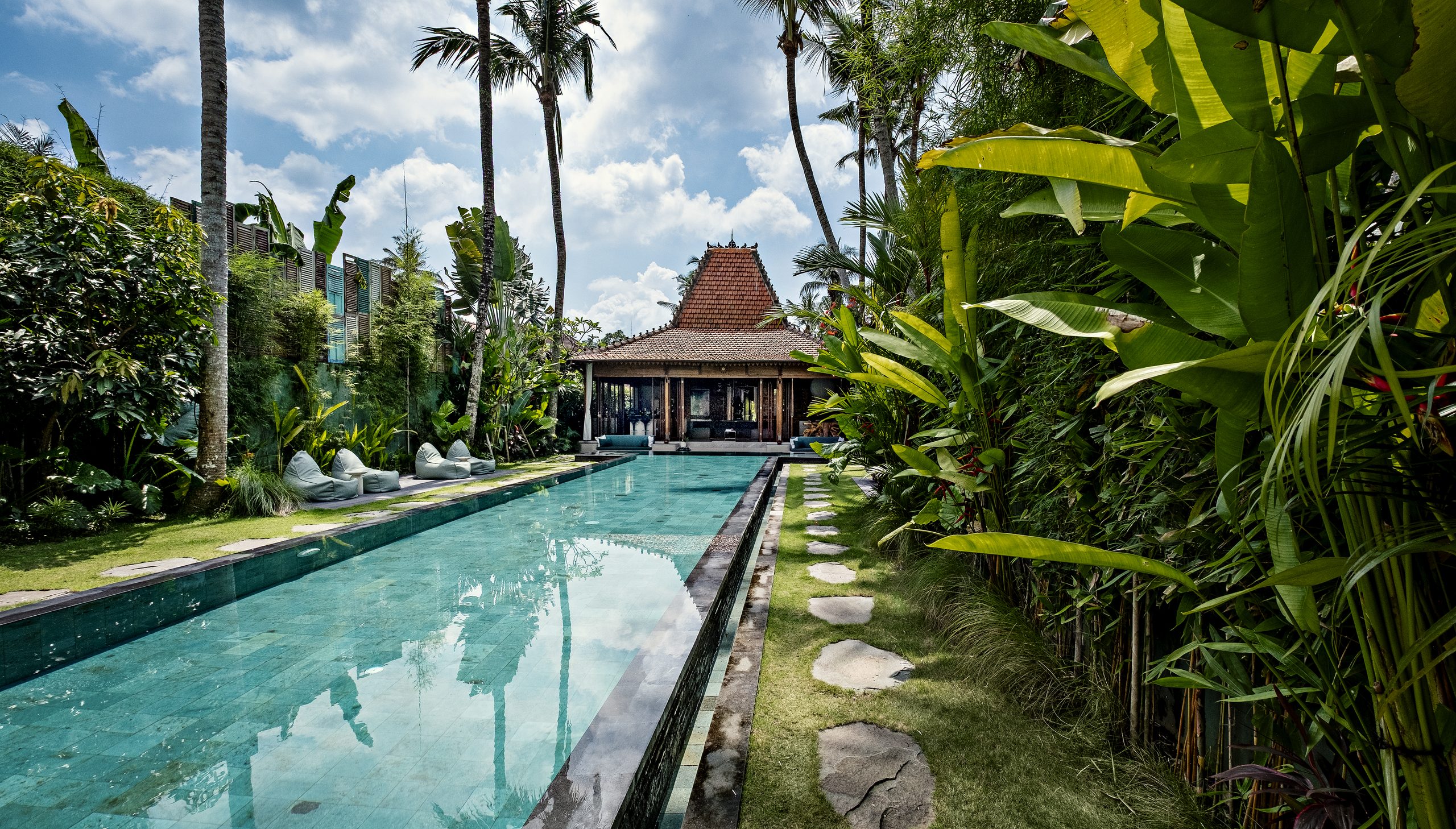 A long turquoise swimming pool surrounded by lush tropical greenery and towering palm trees leads to a traditional Balinese-style pavilion beneath a bright blue sky.