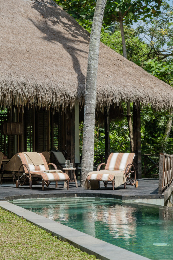 Two cushioned lounge chairs sit beside a turquoise pool and a thatched-roof pavilion surrounded by lush tropical greenery and palm trees.