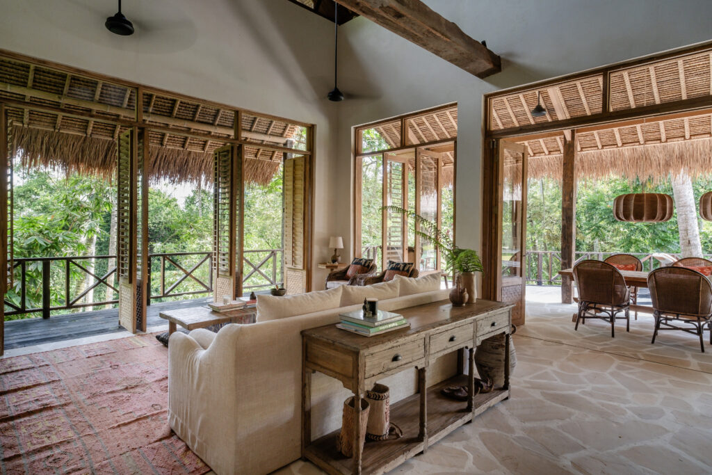 An airy tropical living room with rustic wood furnishings and exposed beams opens through large shuttered doors to a covered dining terrace surrounded by lush greenery.