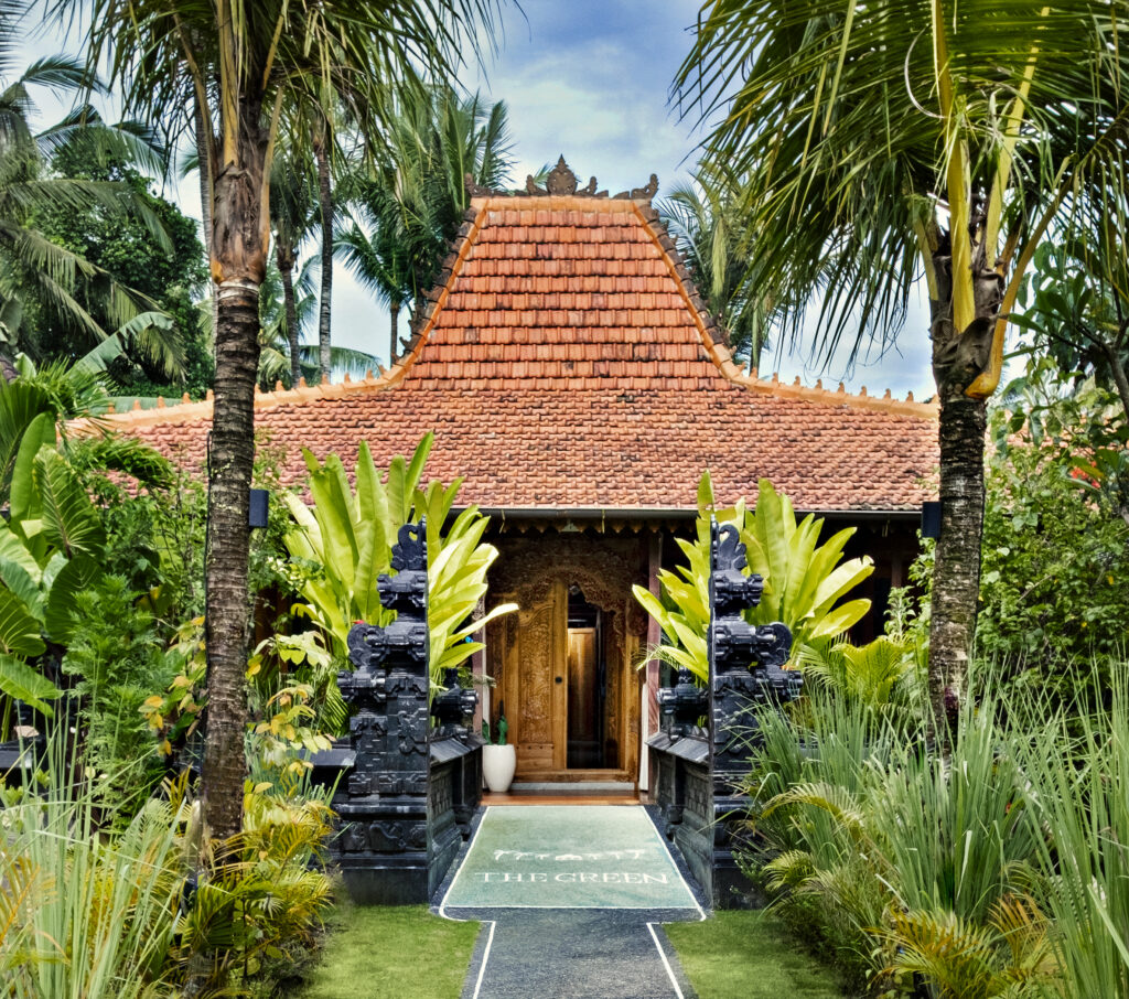 A traditional Balinese-style villa entrance with an ornate red-tiled roof and carved stone gateway is framed by towering palms and lush tropical landscaping.